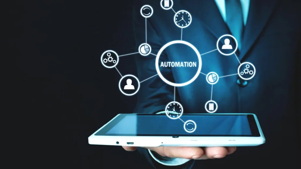 The image depicts a person in a suit holding a tablet. From the tablet, a graphic interface emerges, displaying icons connected by lines. The central theme of the image revolves around the word “AUTOMATION.” This visual representation highlights the integration of automation technology into handheld devices, emphasizing its importance for the success of a passive income business