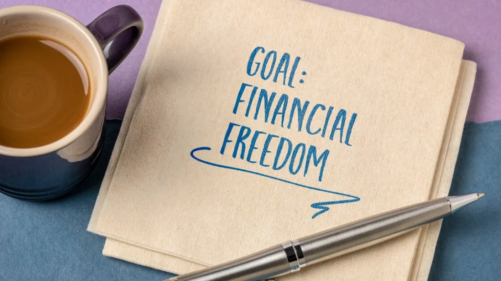 A cup of coffee, a notebook with ‘GOAL: FINANCIAL FREEDOM’ written on it, and a pen are laid out on a surface, symbolizing the planning and motivation required to achieve financial independence.